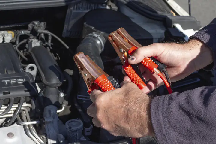 Dłonie mechanika trzymające przewód do łączenia akumulatorów samochodowych, w tle akumulator w silniku